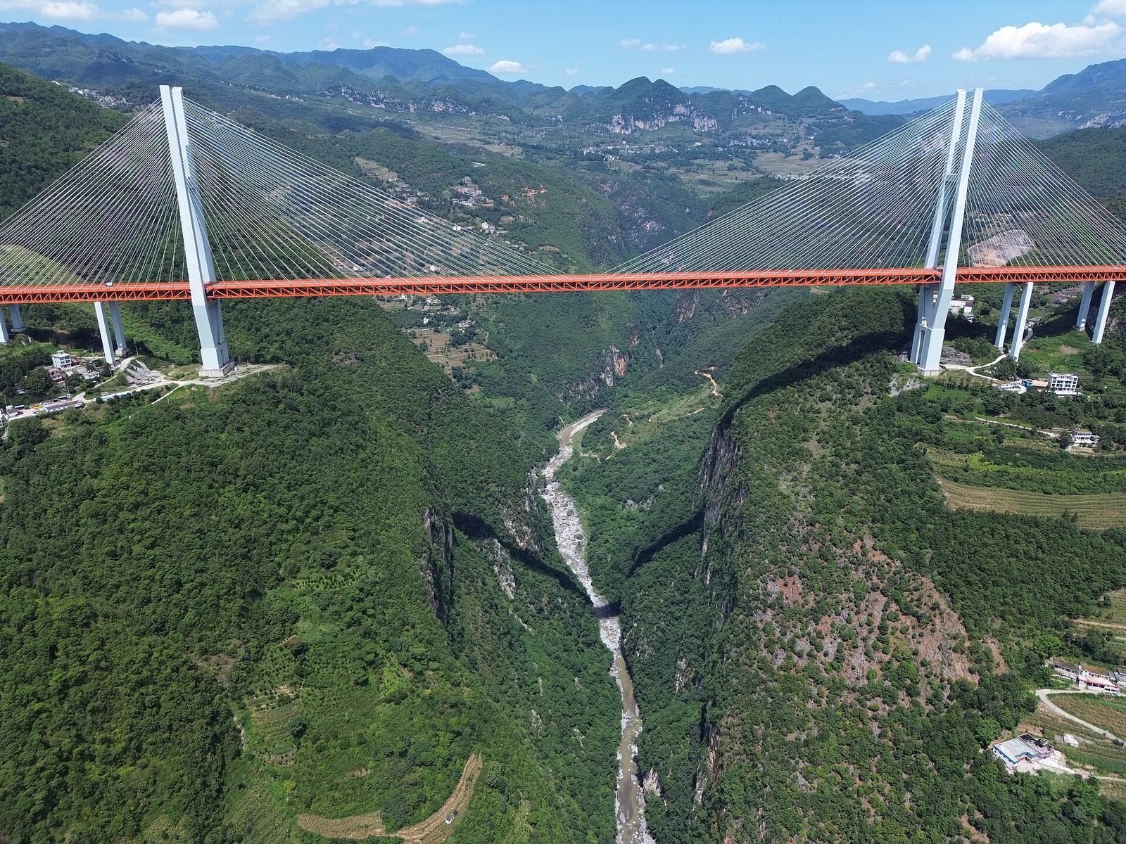 Aerial view of a cable-stayed bridge with a red truss girder between two white towers, passing far above a green gorge, with a narrow river at the bottom