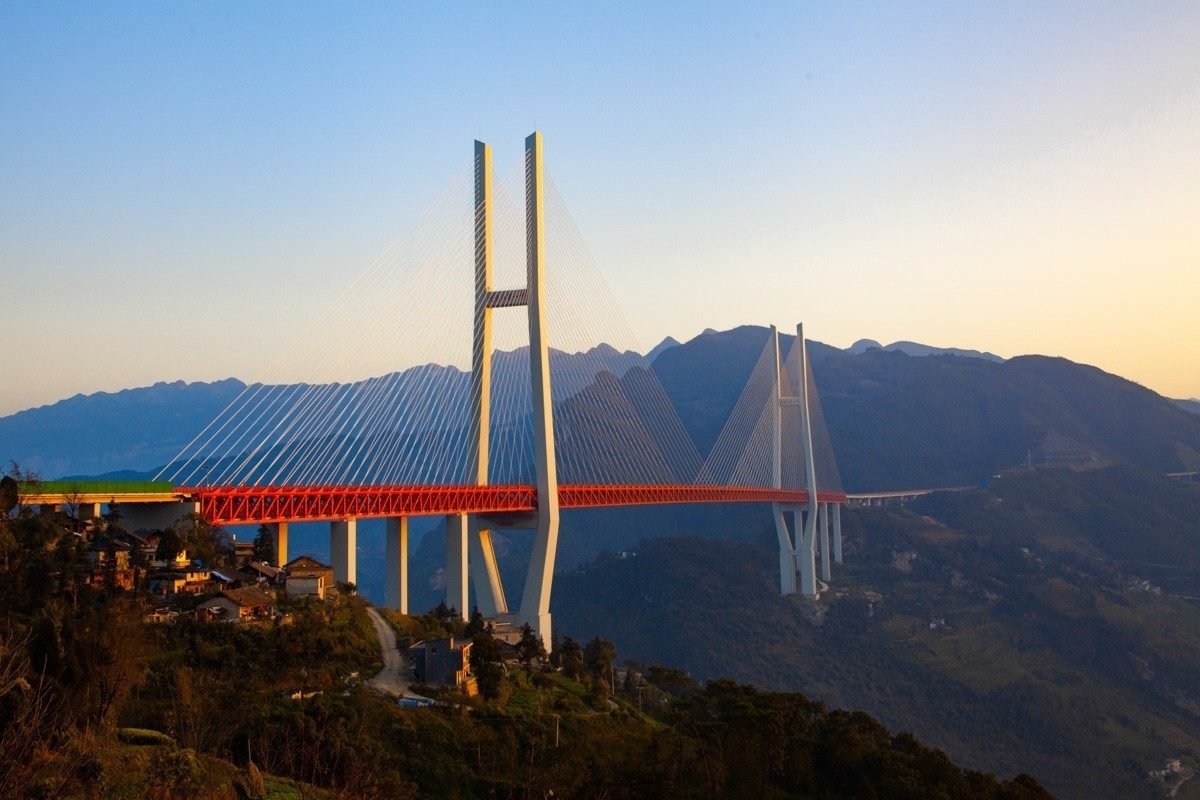 Ponte estaiada de torres brancas e viga vermelha iluminada pelo sol baixo, com montanhas azuladas ao fundo e casas na encosta em primeiro plano