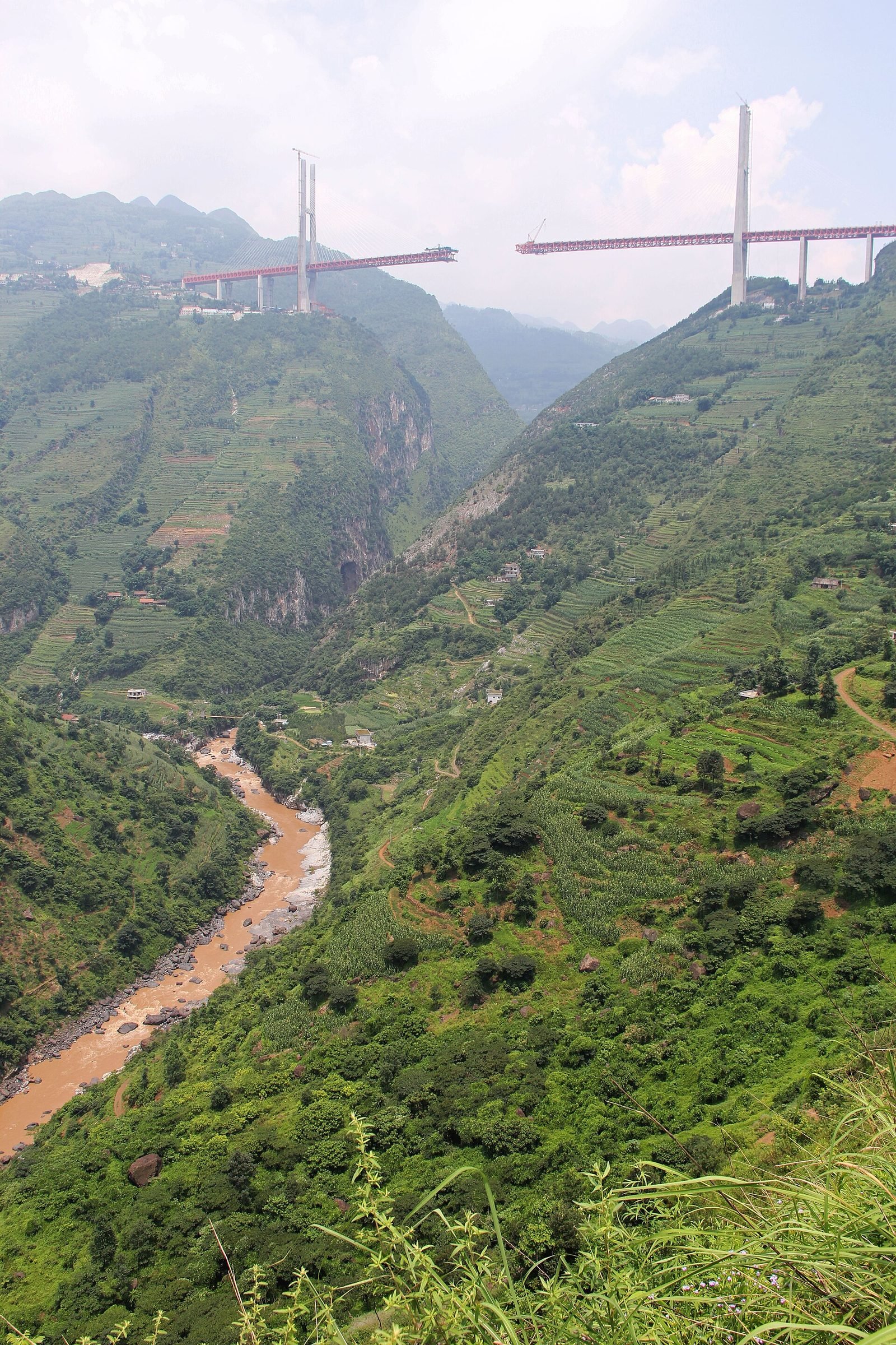 Foto vertical de um vale profundo com um rio barrento no fundo e, lá no alto, as duas torres e os braços de treliça da ponte em construção