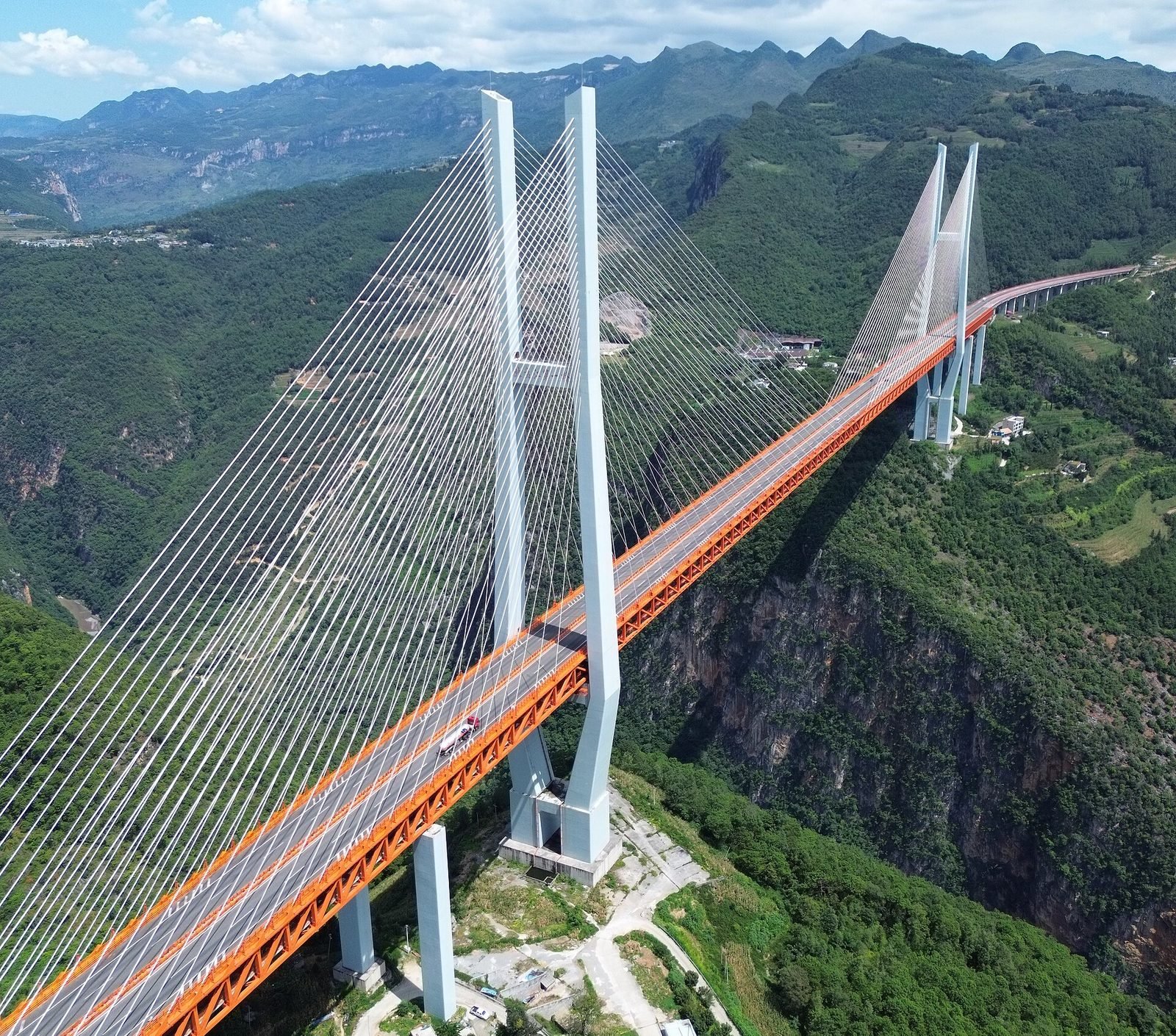 Vista aérea próxima de uma torre de concreto em forma de H com leques de cabos brancos presos à viga de treliça laranja, ao lado de um paredão de rocha