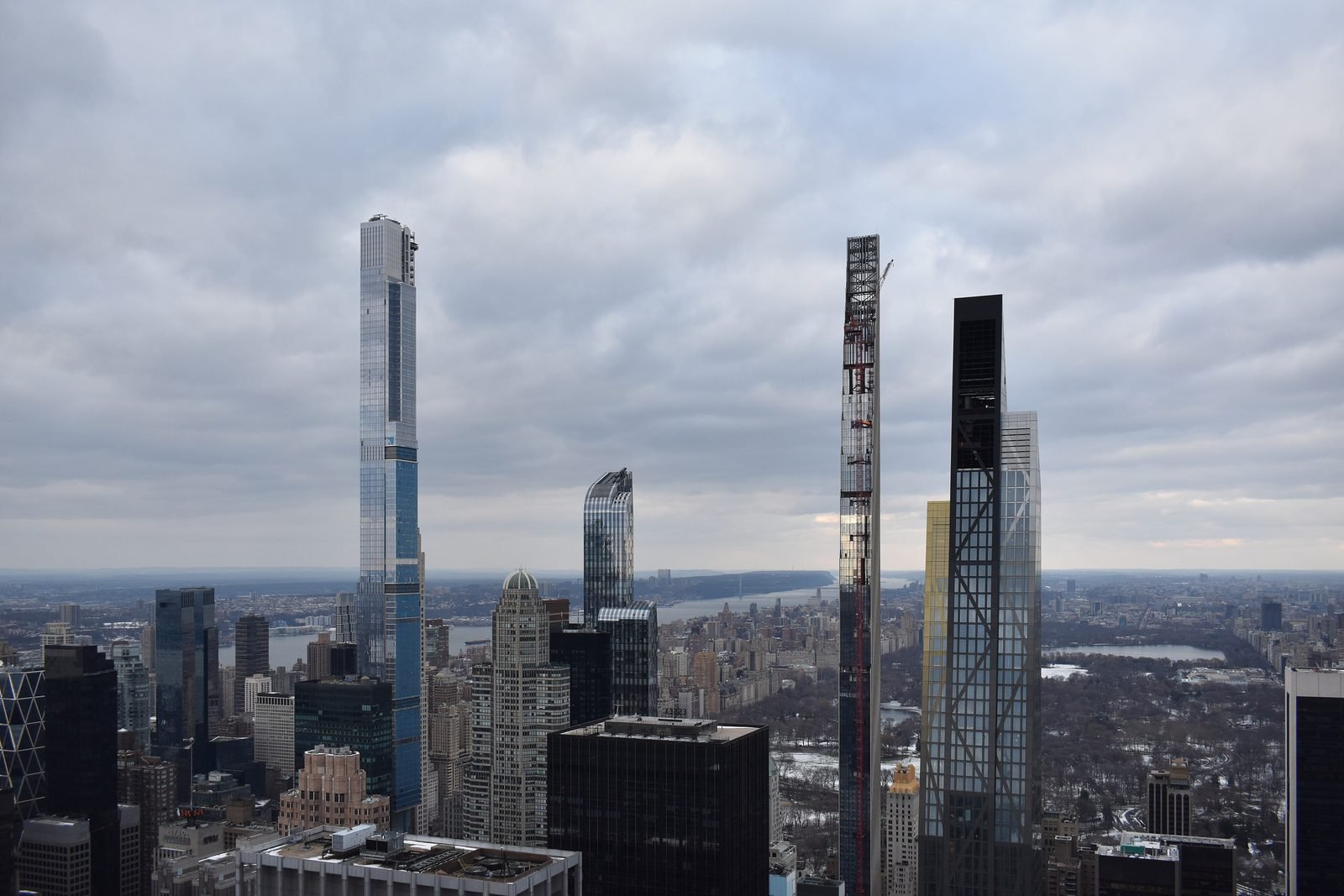 Aerial view of Midtown with several tall, thin towers under a gray sky; on the left the tallest, glass-clad, and on the right an even narrower tower with a crane, with snowy Central Park in the background