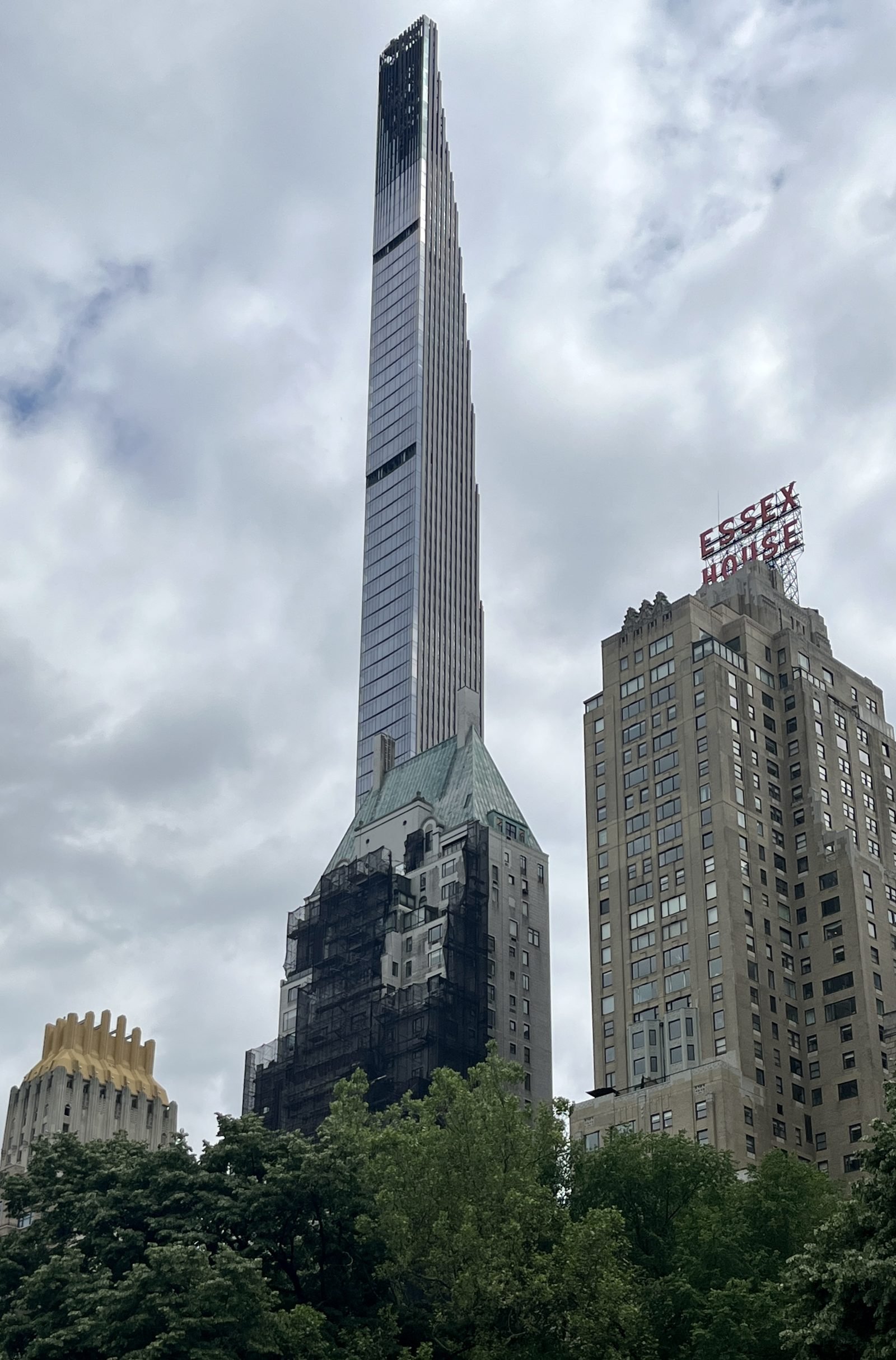 Very thin, tall tower with glass faces that narrow in steps up to the tip, rising above the trees of Central Park under a cloudy sky, with the Essex House hotel sign on a building to the right
