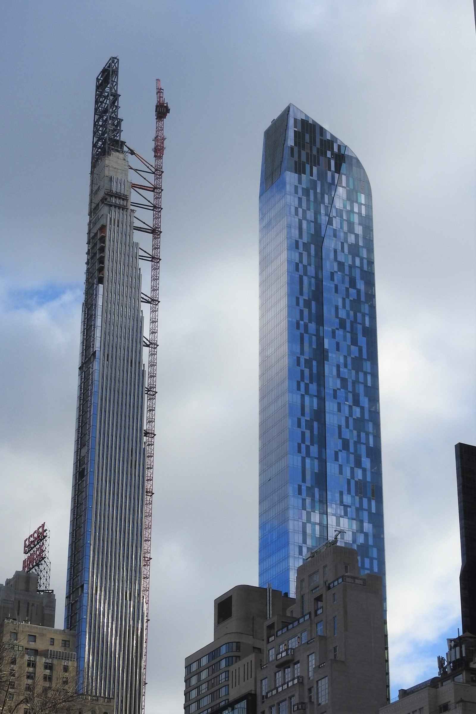 Thin tower under construction with a red crane attached to its side and the top structure still exposed, next to a wider blue glass skyscraper