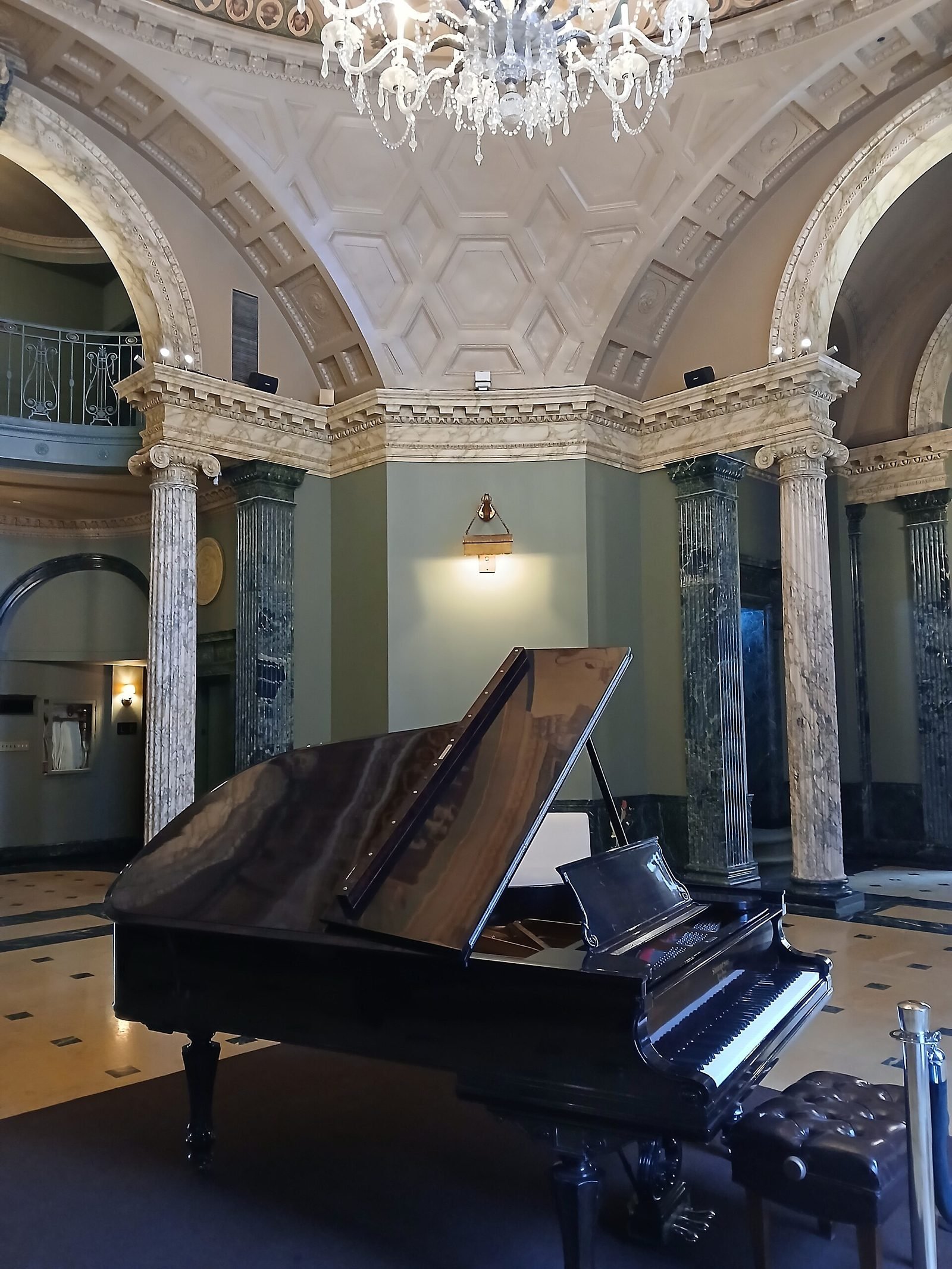 Circular hall with green marble columns, arches, a decorated dome and a crystal chandelier, with a black grand piano in the center