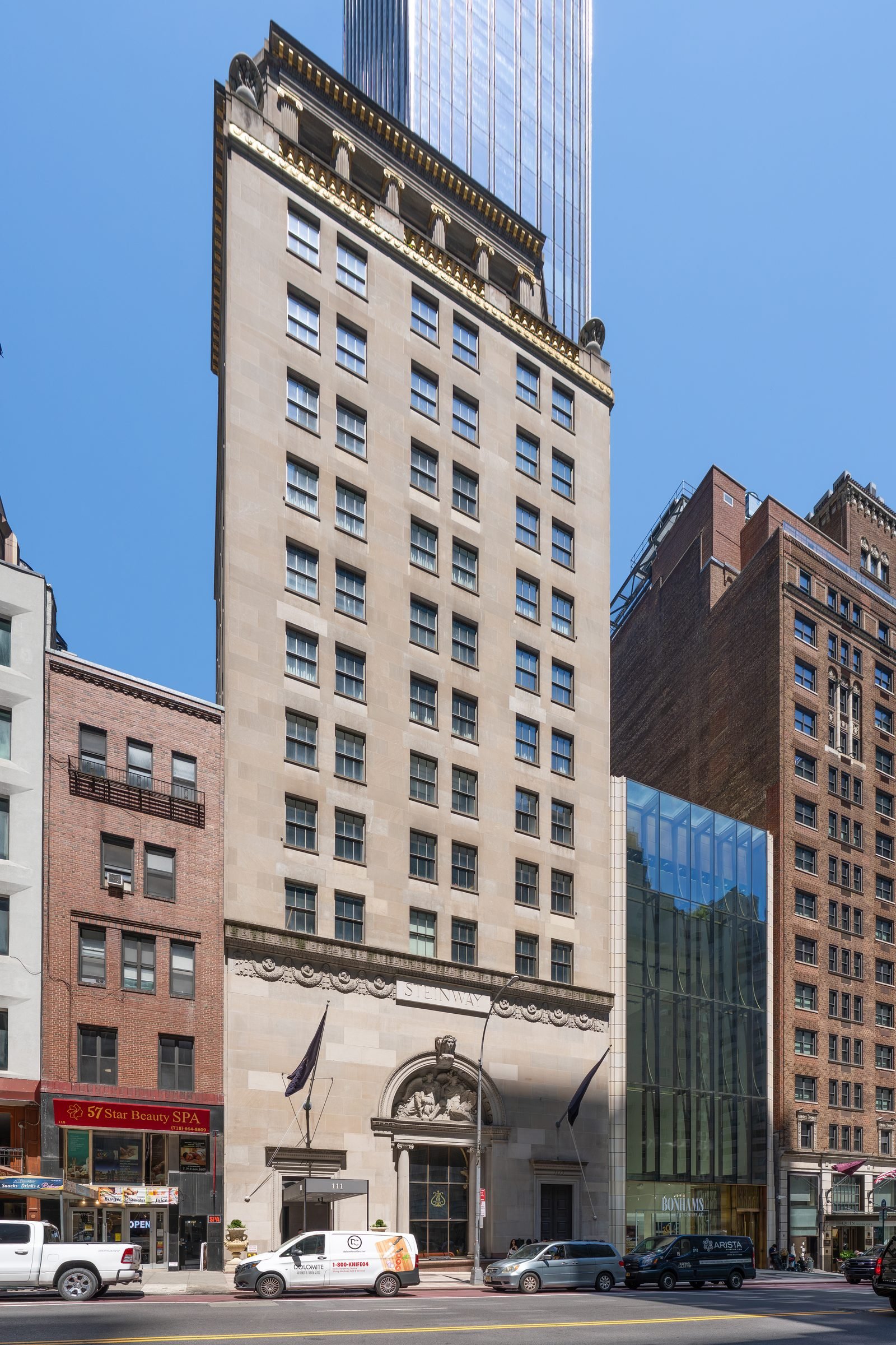Pale stone building several stories high with an arched entrance on the ground floor, on a street with parked cars, and the glass tower rising behind it